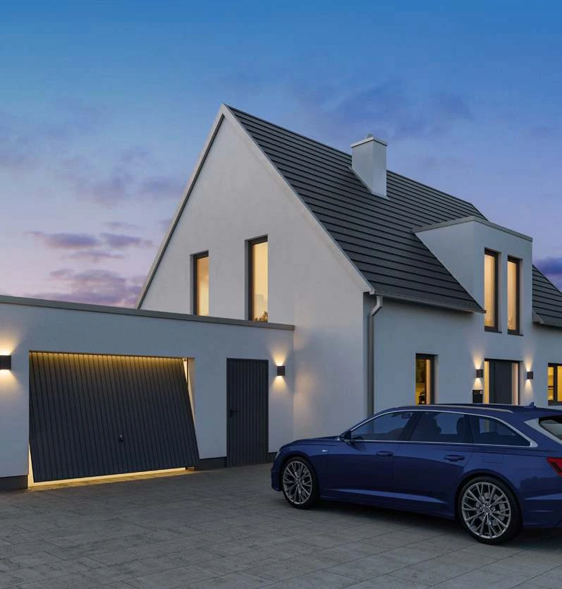 Anthrazitfarbenes Berry-Schwingtor mit senkrechter Rippung an einer Garage neben einem Einfamilienhaus in der Abenddämmerung